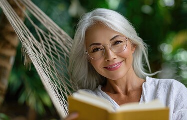 a happy senior woman wearing glasses, sitting in a hammock and reading a book