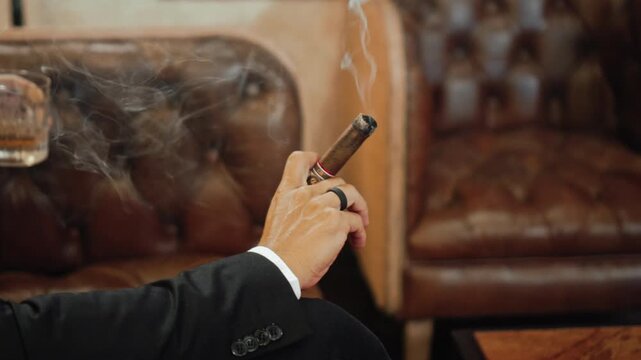 Men's hands hold a smoking cigar while sitting on a leather sofa with a glass of whiskey.