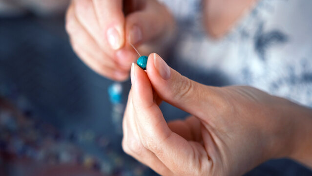 A man makes a bracelet out of blue beads. A close-up of a woman's hands stringing a bead to a bracelet with a thin thread. Hobbies and creativity