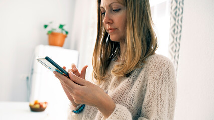A woman is chatting enthusiastically on her phone. The blonde stands in the kitchen and looks at her mobile phone