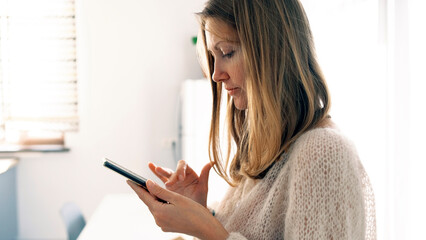 Fototapeta premium The student is looking for something on her phone. A pretty young woman stands in a room and looks at her mobile phone