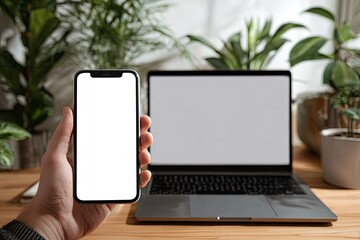Hand holding smartphone with blank screen next to laptop on wooden desk, surrounded by houseplants