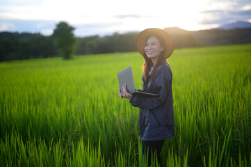 A smiling woman in a hat stands in a lush green field, holding a laptop, enjoying the serene...