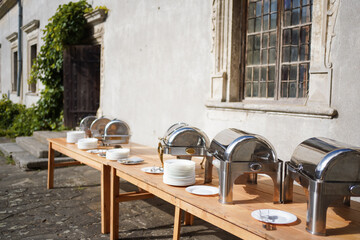 Outdoor buffet setup with stainless steel chafing dishes and stacked plates on a wooden table near an old stone building with large windows.