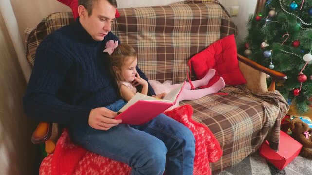 father and little daughter sit on sofa near Christmas tree at home, and read fairy tale book together, smile and laugh at funny stories