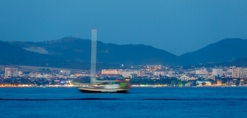 Fototapeta premium Gelecik resort in evening twilight. Pleasure vessel moving along Gelendzhik Bay. City lights and Caucasus Mountains in background.