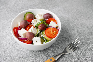 Greek salad with feta cheese olives cucumbers tomatoes onions in white bowl with fork on light grey background