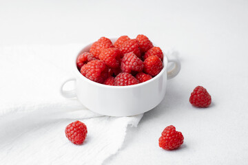 Raspberries in white bowl with scattered berries on light surface for organic food or vegan diet concept