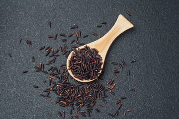 Wooden spoon filled with black rice and spilled grains on dark background