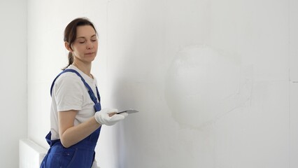 Female construction worker spreading finishing putty on white wall, wearing protective gloves and...