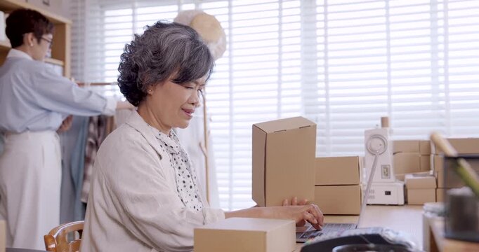 Senior asian woman working laptop while stacking cardboard boxes boutique fashion studio small family business ecommerce entrepreneurship packaging teamwork handmade textile clothing online shop