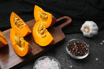 Orange pumpkin pieces with seeds on cutting board, garlic, peppercorns and salt, dark background