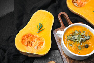 Butternut squash soup with pumpkin seeds and rosemary on wooden board, dark fabric background.