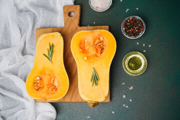 Halved butternut squash with rosemary on wooden board, salt, peppercorns, oil on table