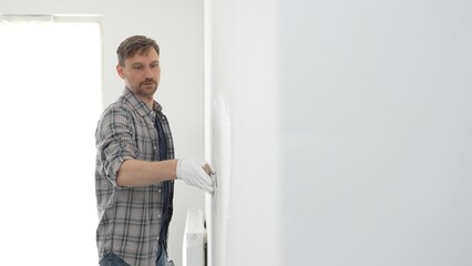 Construction worker wearing gloves and using a putty knife to plaster a white wall, performing home...