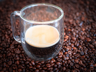 Glass cup with espresso coffee placed on roasted beans. Fresh hot drink with crema foam, symbol of morning energy, relaxation and caffeine boost.
