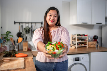 Portrait of Asian plus-size woman holding a bowl of salad in kitchen at home. 
