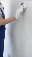Close-up of man construction worker wearing gloves and blue overalls is using a putty knife to...