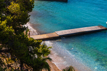 The Aegean coast on a sunny summer day in Kusadasi. Azure water, piers, green hills, palm trees and a promenade with the flag of Turkey. A picturesque seascape, a popular tourist destination for