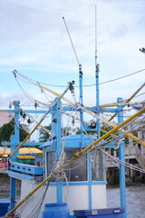 A blue and yellow fishing boat docked at the pier, equipped with tall poles and fishing gear, set against the bright seaside background.