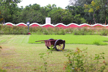 Two-wheeled handcart on a freshly mown lawn with a red wavy wall and trees beyond. Quiet daylight scene for gardening, park maintenance, agriculture and rural lifestyle projects.