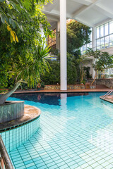 Luxury indoor swimming pool surrounded by exotic tropical plants and modern glass architecture. Relaxing wellness environment with natural light, reflections on the water and resort atmosphere