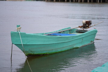 Naklejka premium A light green long-tail boat with an old engine floats calmly on the water, secured with a rope to the dock.
