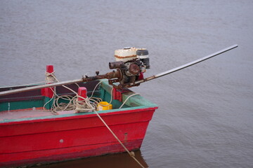 A red long-tail boat floating on calm seawater under a partly cloudy sky, with distant islands on the horizon, capturing the peaceful simplicity of coastal fisherman life