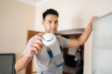 Selective focus portrait of blurred electrician holding smoke detector while installing in residential building, prioritizing fire safety and ensuring protection for all occupants within home.