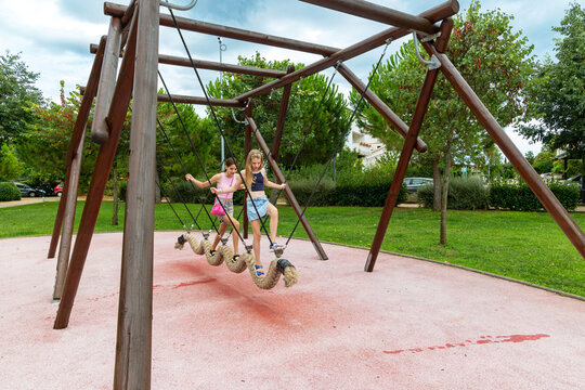 Teen girls balancing on rope swing in park. Two teenage girls play and balance on a wavy rope swing in a green public park, enjoying an active outdoor afternoon together.