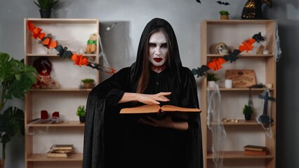 A witch or vampire woman in a black cloak reads a spell book on Halloween