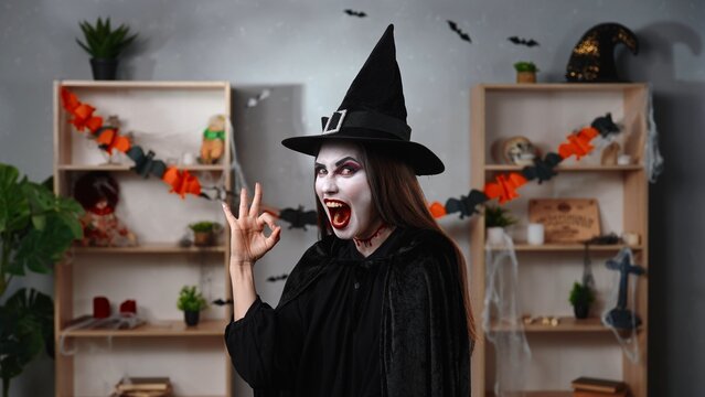A vampire witch in a black cloak and hat makes an OK gesture with her fingers