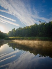 Morning at a picturesque pond in Bohemian Paradise. Above the water, steams illuminated by the sun's rays. Greenery all around.
