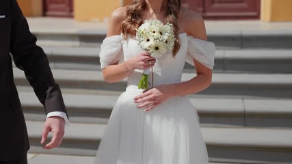 Bride in elegant strapless gown holds a white floral bouquet, standing outside the venue