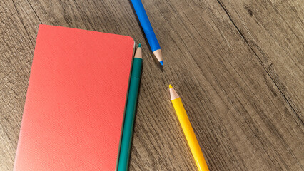 A composition of a blank red notebook and three colorful pencils on a textured wooden background.