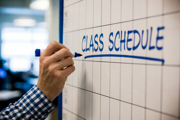 Hand writing C L A S C H E D U L E on a whiteboard with blue marker class schedule