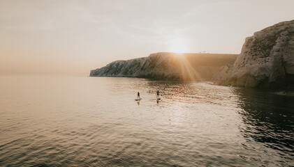 Two people paddleboards on a calm ocean near a rocky shore. The sun is setting, casting a warm glow over the water. The scene is peaceful and serene, with the sound of the waves