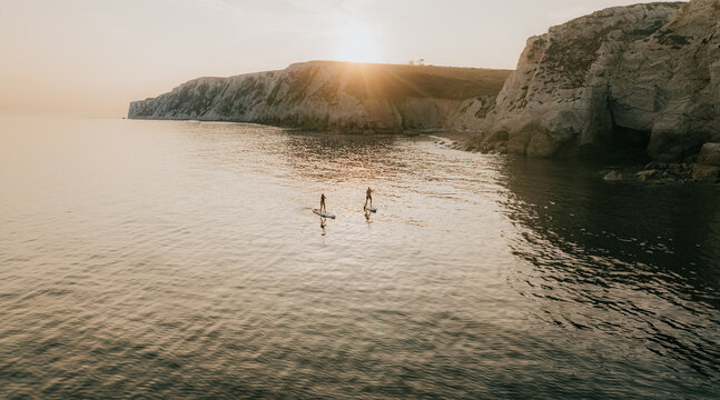 Two people paddleboards on a calm ocean near a rocky shore. The sun is setting, casting a warm glow over the scene