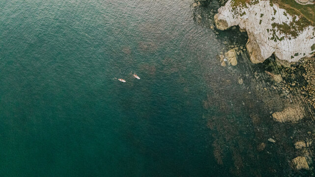 Two people paddle in the ocean near a rocky shore. The water is calm and the sky is clear - Powered by Adobe