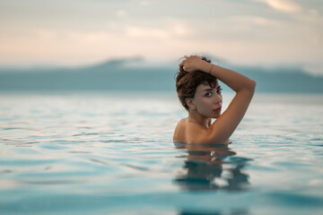 beautiful woman swimming in luxury pool at sunset flowing into sea, elegant vacation and tropical serenity, stunning girl relaxing in lavish resort pool at sunrise