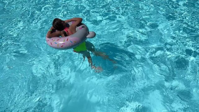 Young girl relaxing on cute sloth inflatable pool float with colorful sprinkles in crystal clear blue swimming pool water - summer vacation concept