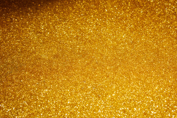 a close-up of a golden, glittery surface. The texture is densely packed with tiny, shimmering particles that reflect light, creating a sparkling effect.