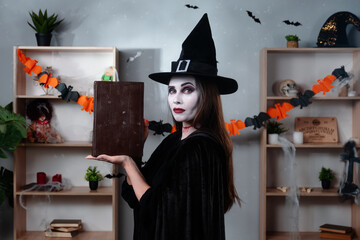 A vampire witch in a black cloak and hat shows off a book of magic while standing in a room...