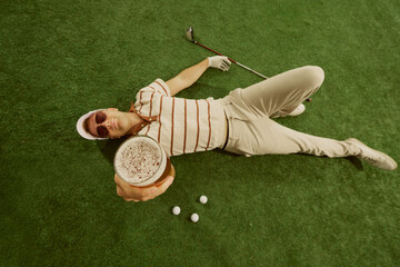 Male golfer lying on grass holding beer glass forward with playful relaxed expression. Concept of lifestyle leisure, beverage culture, sports advertising and relaxation branding.