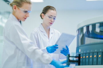 A senior scientist supervises a junior lab technician operating an automated analyzer. Two female...