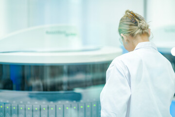 A female laboratory technologist monitors an automated clinical analyzer processing patient samples. Green status lights indicate the active diagnostic workflow in the high-tech lab.
