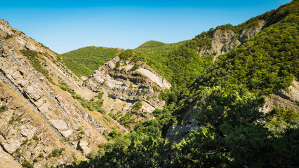 Fototapeta premium Geological layers of folded rock formations rise among green hills near Armazi Fortress in Mtskheta, Georgia. A dramatic view of natural power and raw Caucasus beauty