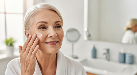 Fototapeta premium Skincare routine for mature skin. Woman applying face cream in bright bathroom. Daily skincare routine includes moisturizing cream for healthy skin.