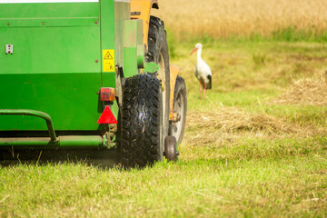 Rear of a hay baler machine with a tractor in a meadow and a standing stork