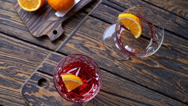 Italian cocktail known as Negroni served in transparent glassware garnished with slice of citrus fruit and accompanied by fresh, uncut oranges on cutting board alongside straining tool, all arranged 